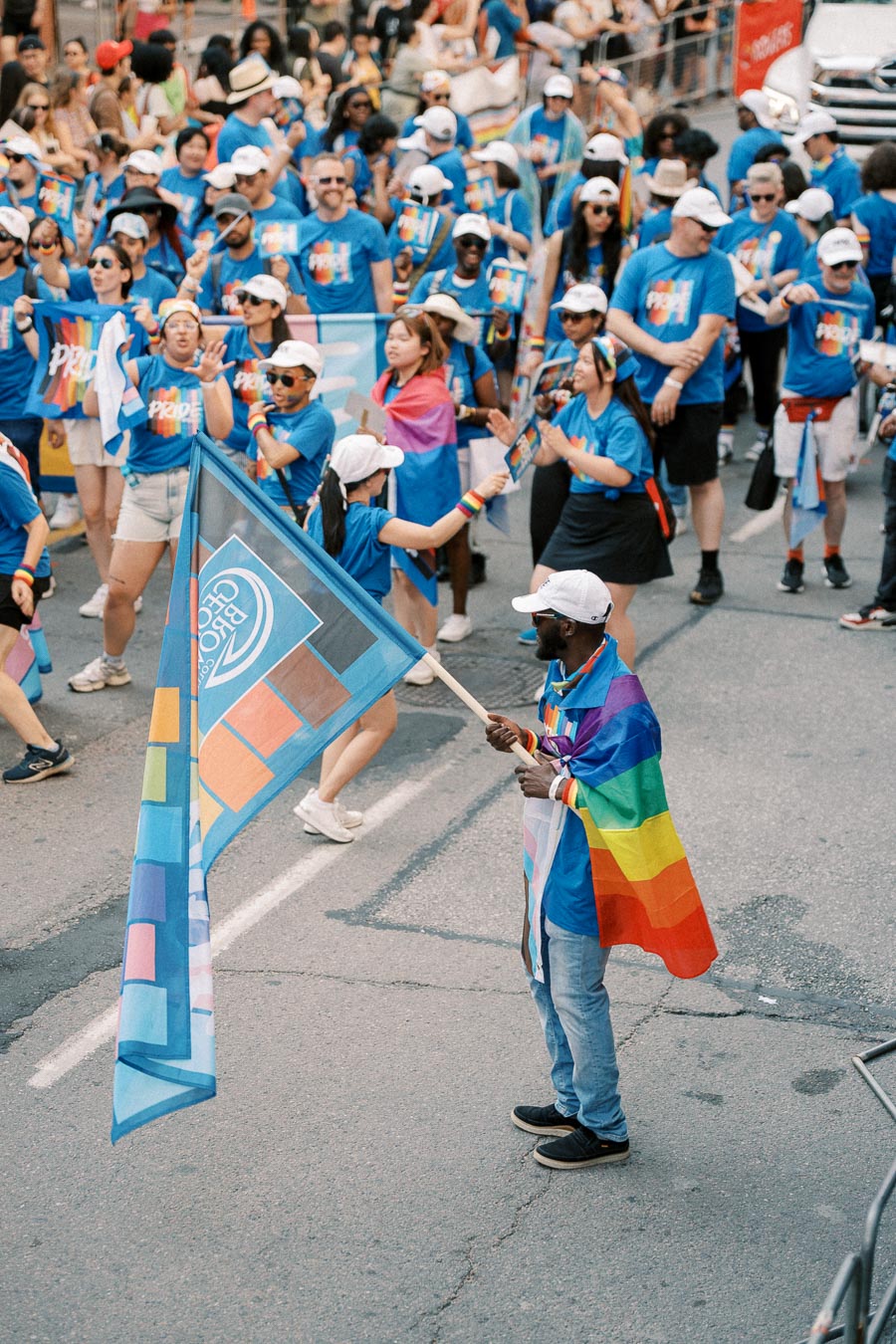 Participants in a Pride parade wearing blue shirts and white caps, carrying rainbow flags and banners.