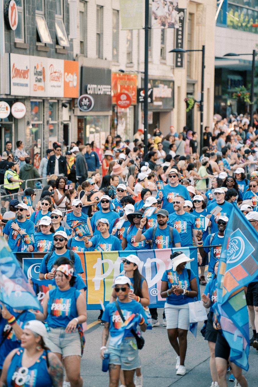 Participants in a Pride parade march down a city street, wearing matching blue shirts and white hats, while holding flags and banners.