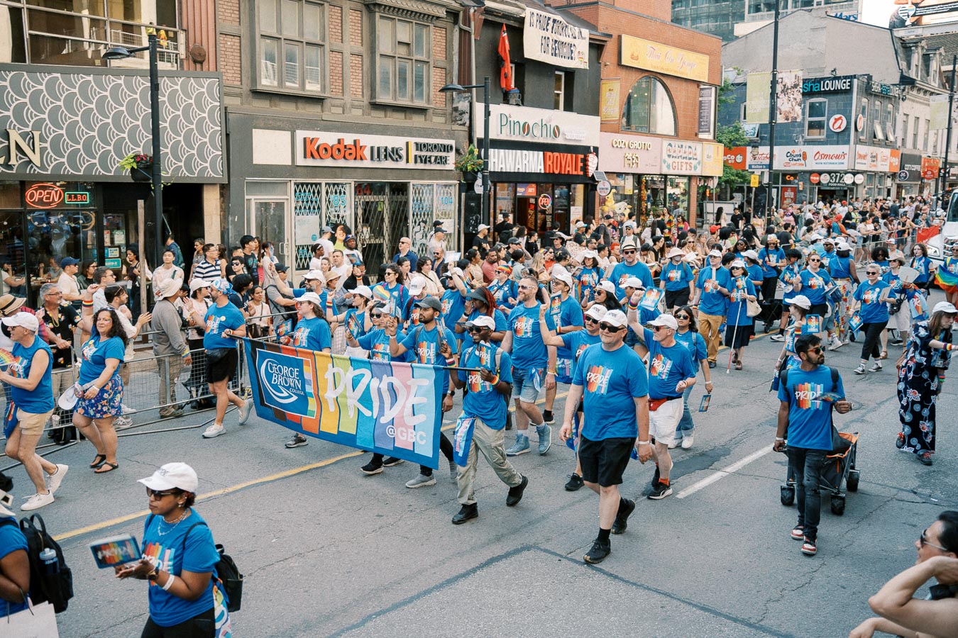 Participants from George Brown College march in a Pride parade, wearing blue shirts and carrying a banner. Spectators line the street, enjoying the festive atmosphere.