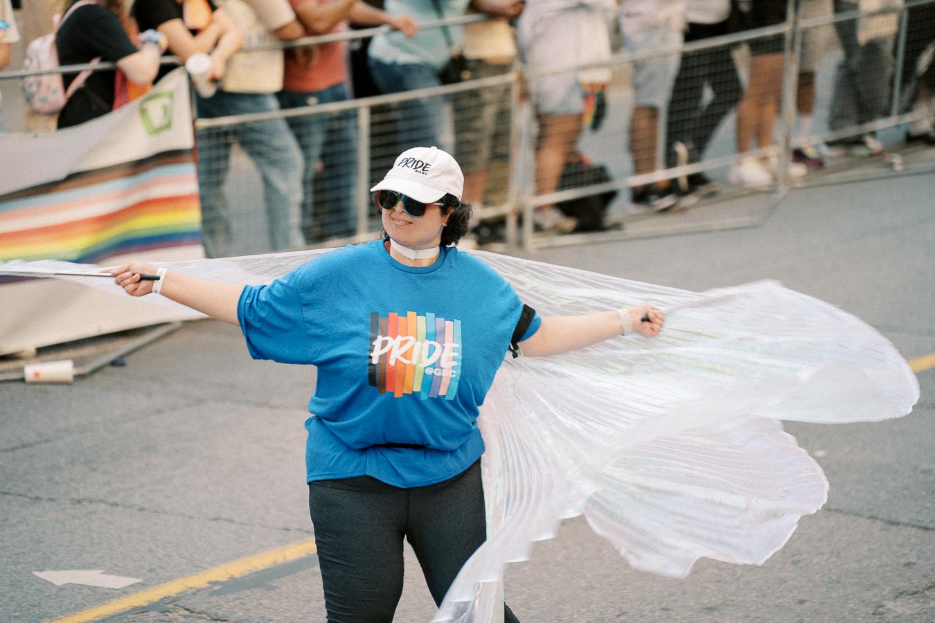 A participant in a Pride parade wearing a blue t-shirt with a rainbow design and the word 'Pride', along with a white cap. They are holding translucent wings, adding a festive touch to the event.