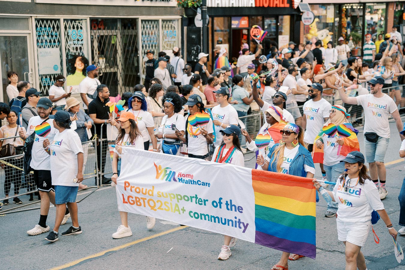 Participants in a Pride parade march with a banner supporting the LGBTQ2SIA+ community, wearing rainbow-themed attire and carrying flags.