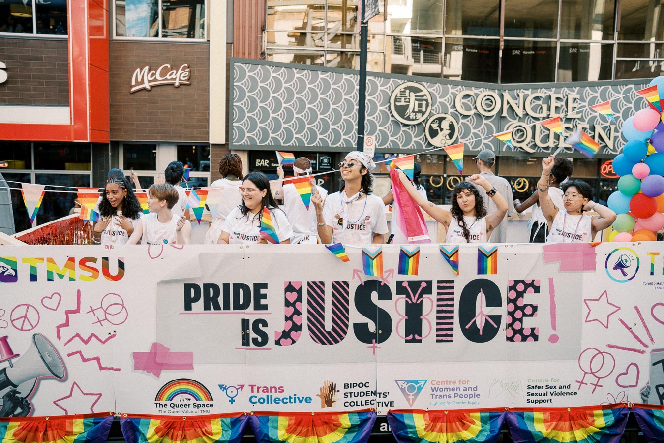 Participants celebrate at a Pride event, holding rainbow flags and standing behind a banner that reads 'Pride is Justice'.