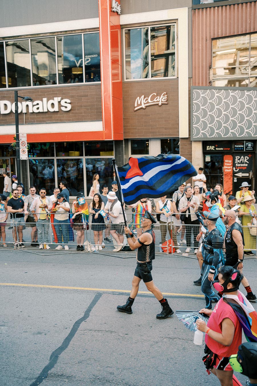 A participant in a leather outfit carries a flag during a pride parade, with spectators watching from behind barriers in front of a McDonald's.