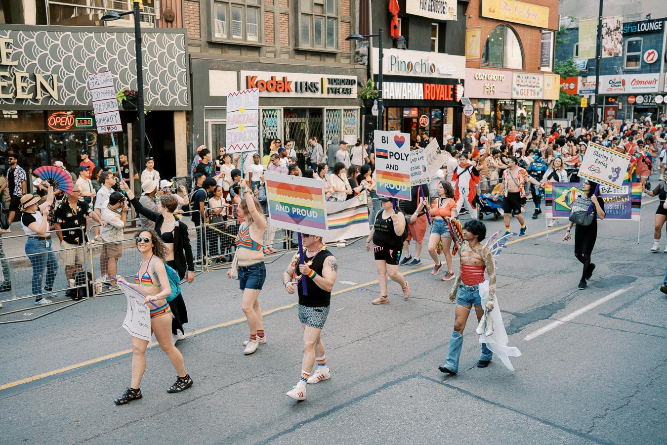 Participants in a pride parade march down a city street, holding signs advocating for polyamory and LGBTQ+ rights, surrounded by a crowd of onlookers.
