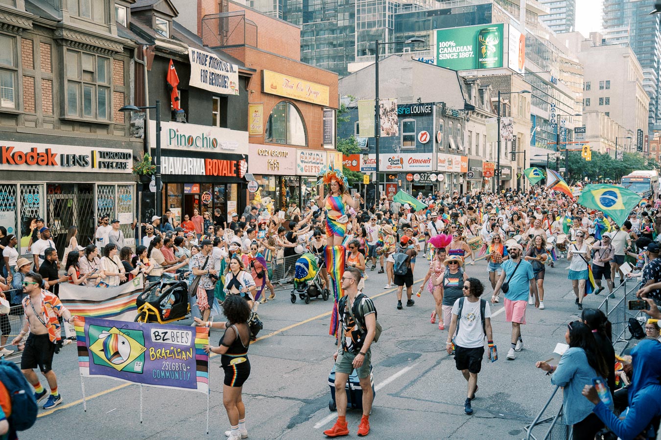 A lively parade featuring participants in colorful costumes and carrying flags, taking place on a bustling city street lined with various shops and spectators.