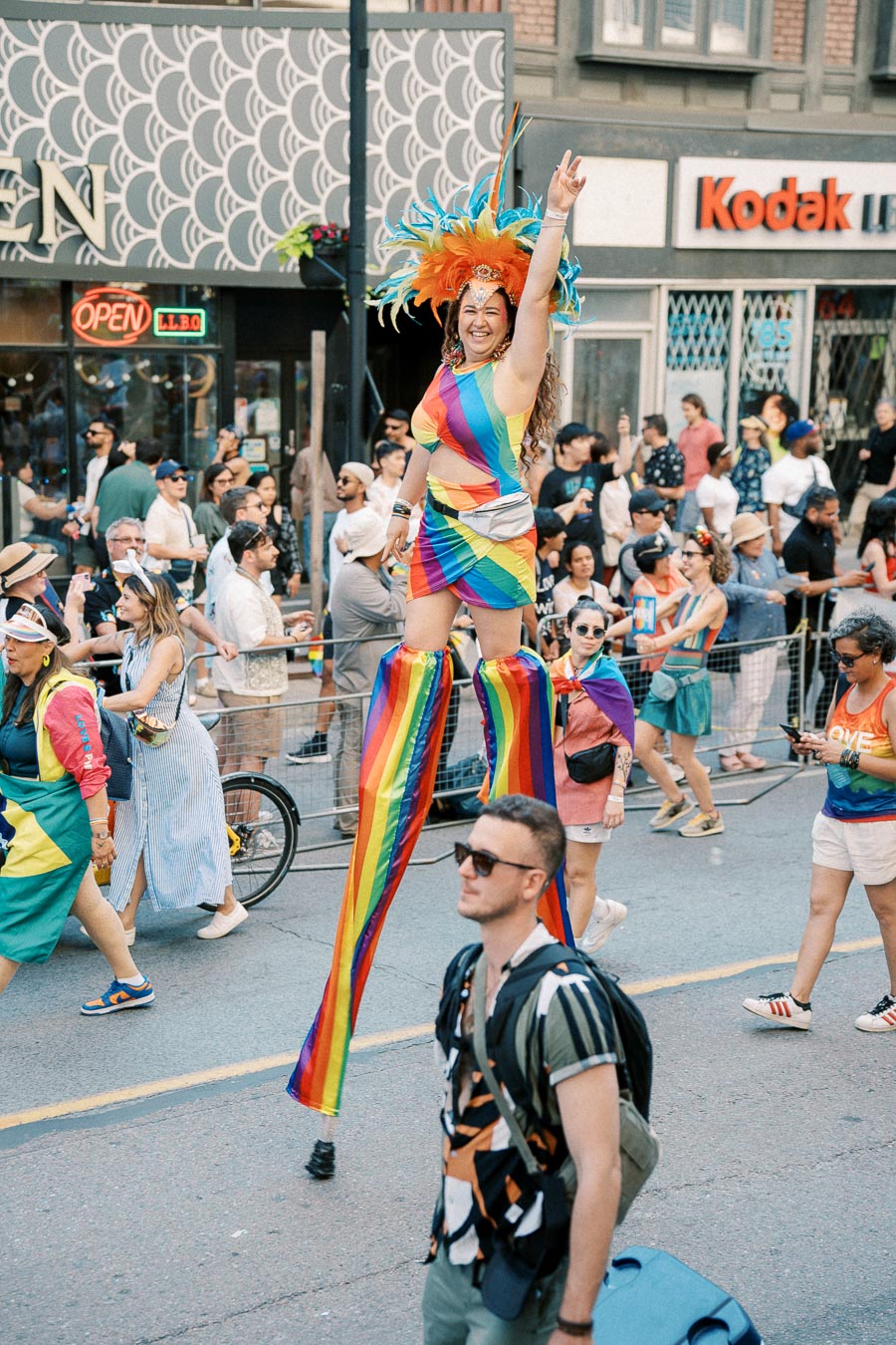 A vibrant scene from a pride parade featuring a person on stilts dressed in a rainbow outfit and feathered headdress, surrounded by a lively crowd.