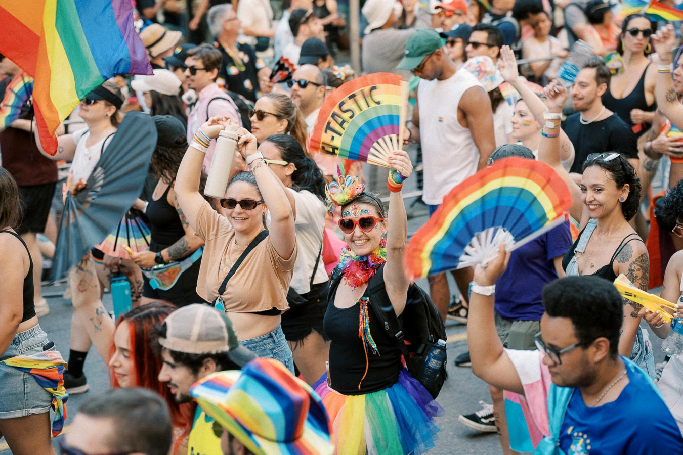 Participants celebrate at a Pride parade, waving rainbow flags and fans, dressed in vibrant, colorful attire.