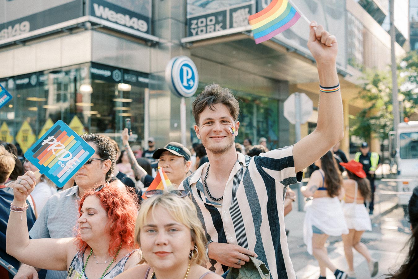 Participants celebrate at a Pride parade, waving rainbow flags and holding signs, surrounded by a lively crowd on a city street.