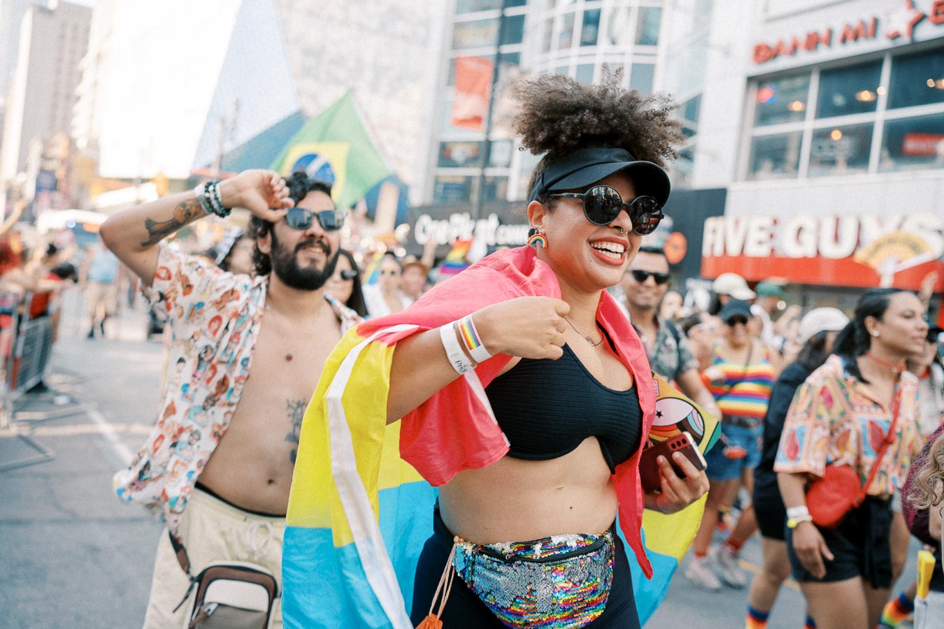 A lively scene from a pride parade featuring participants in colorful outfits and flags, including a Brazilian flag. The background shows a bustling city street with recognizable storefronts like Five Guys.