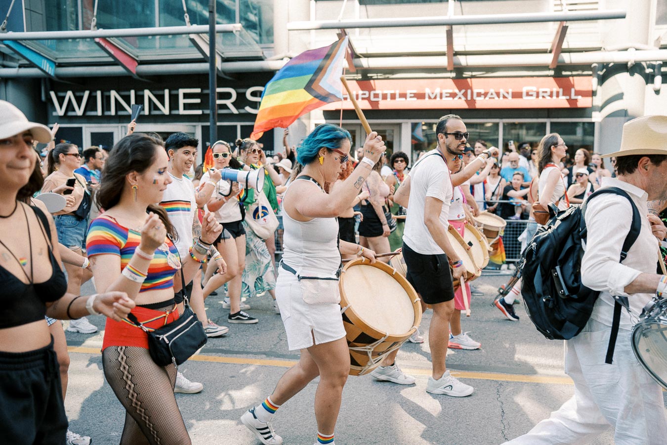 Participants in a pride parade march with a rainbow flag and drums, celebrating diversity and inclusion.