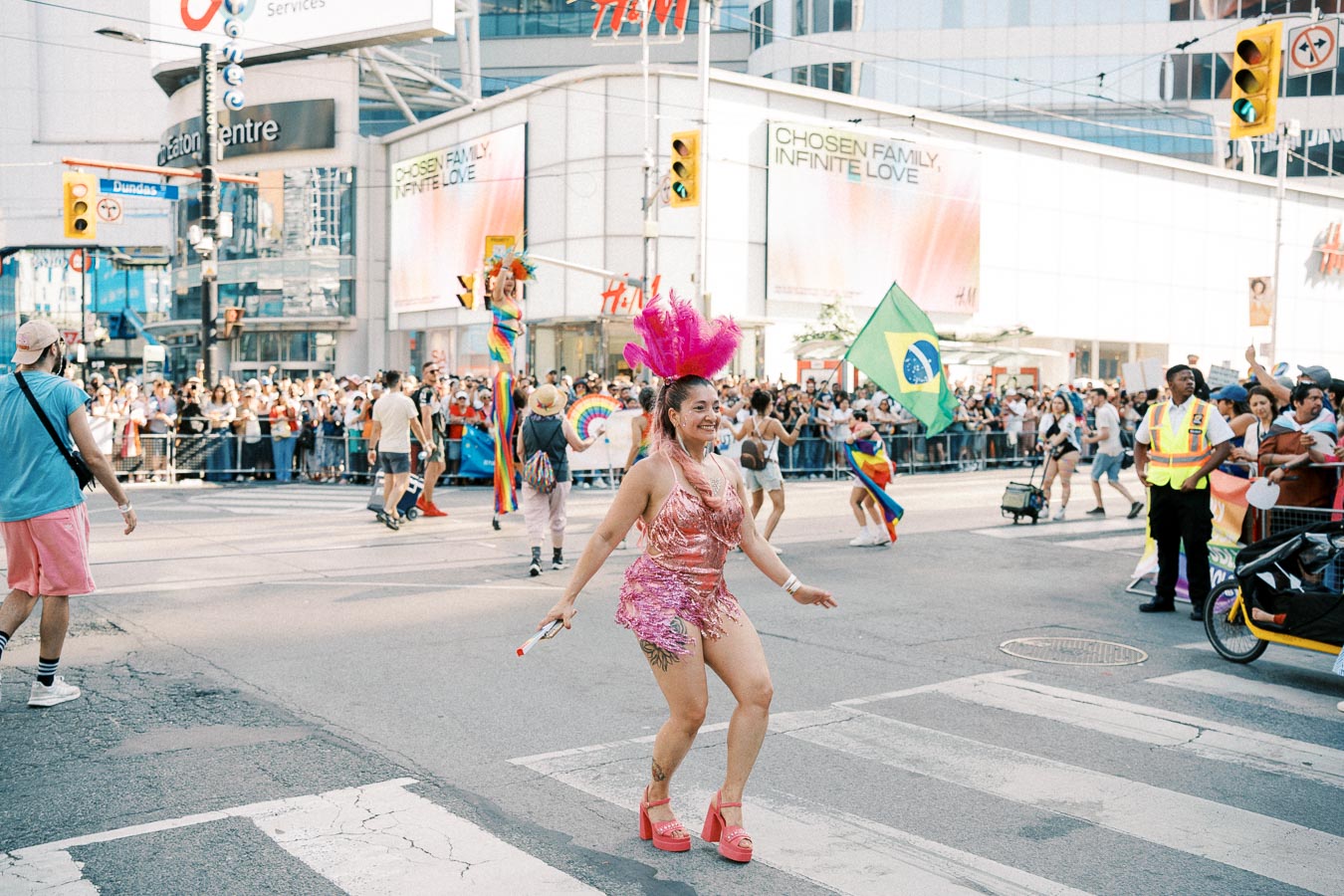 A lively parade featuring a dancer in a pink costume and feathered headpiece, with participants waving a Brazilian flag and rainbow flags, set against a bustling city backdrop.