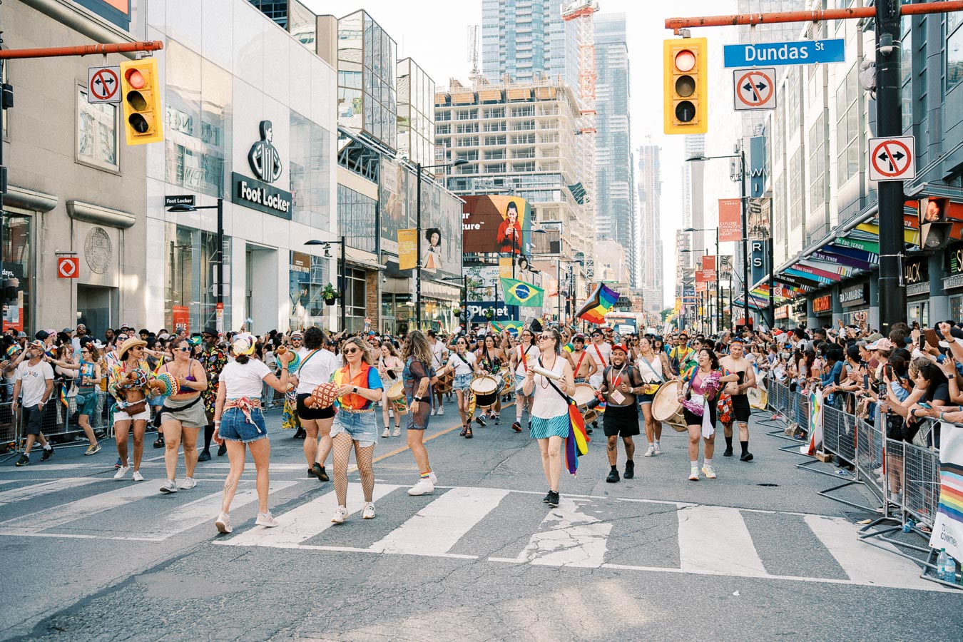 A lively parade takes place on Dundas Street, featuring participants with musical instruments and rainbow flags, celebrating diversity and inclusion.