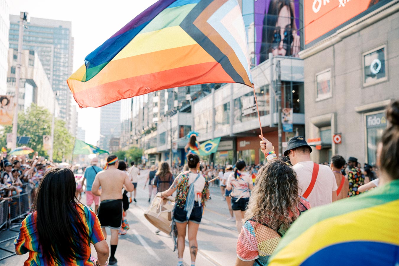 Participants march in a pride parade, waving a rainbow flag symbolizing inclusivity and diversity, surrounded by cheering spectators and city buildings.