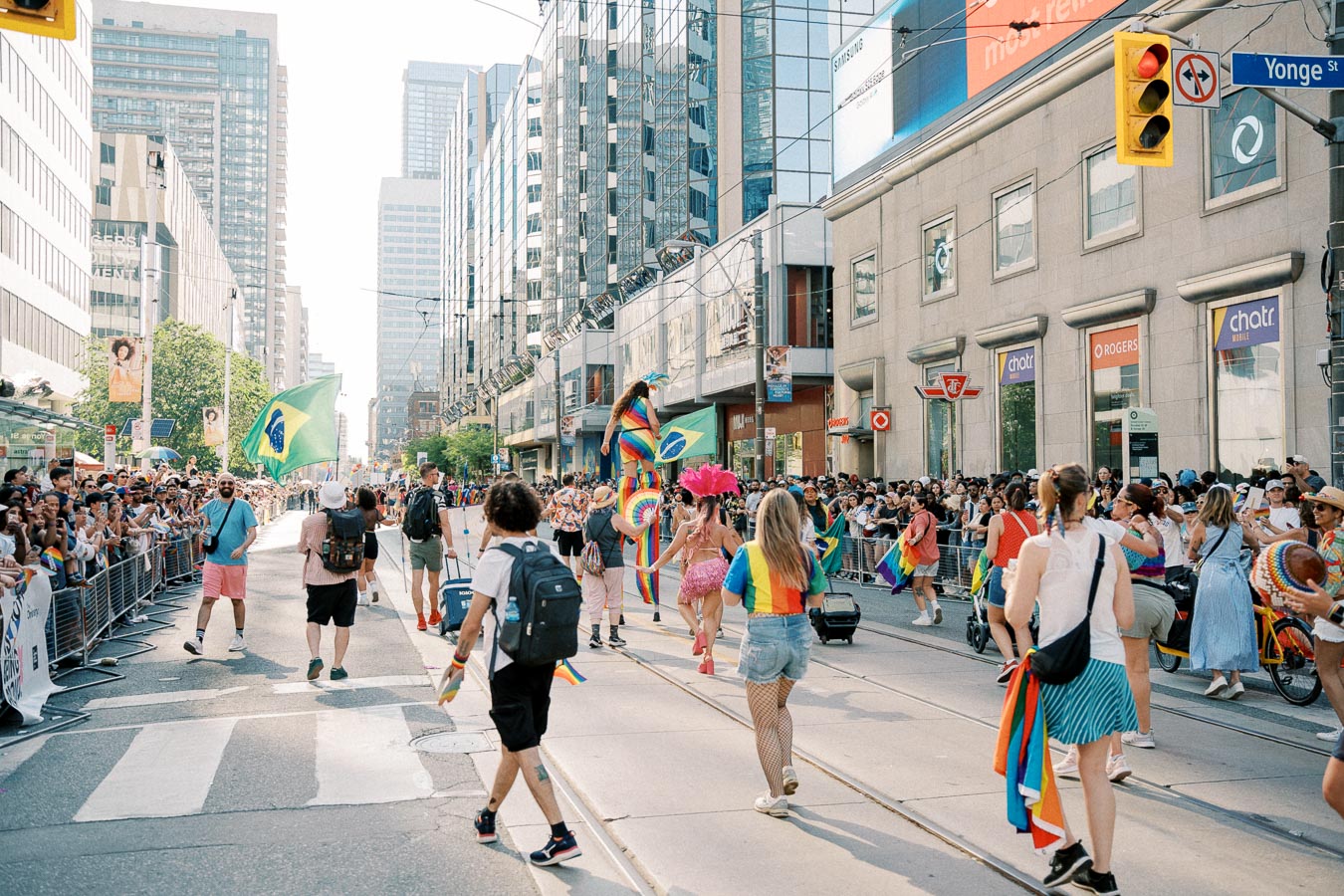 A lively parade takes place on a bustling city street, featuring participants with rainbow and Brazilian flags, surrounded by cheering crowds and tall buildings.