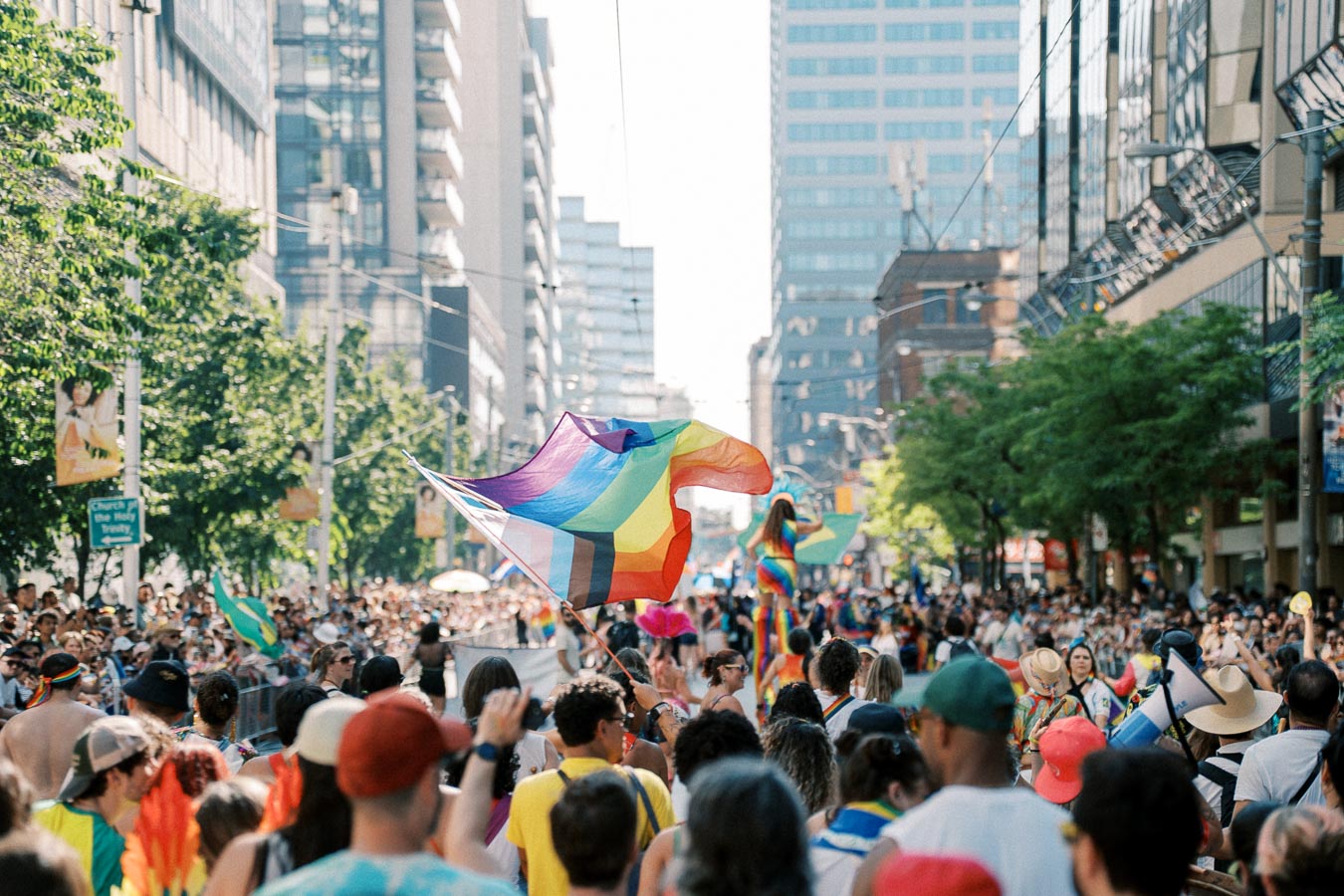 A lively pride parade with a large crowd, rainbow flags, and colorful outfits, set in an urban environment.