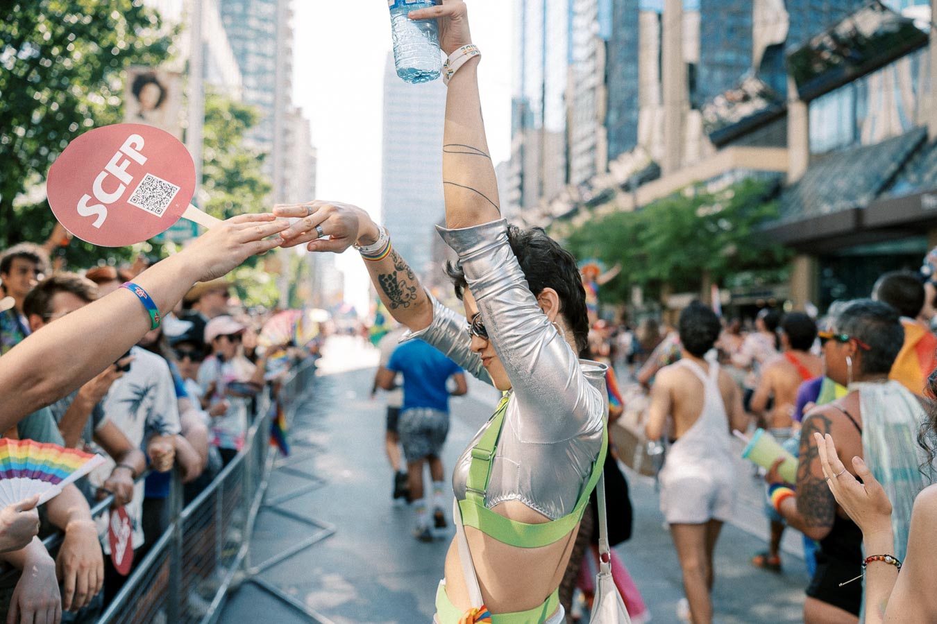 A lively scene at a parade with a person in a silver and green outfit dancing, while the crowd cheers and holds colorful fans and signs.