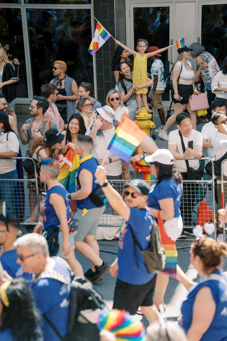 A vibrant scene from a pride parade, featuring a diverse crowd celebrating with rainbow flags. A young girl in a yellow dress stands on a fire hydrant, joyfully waving a flag.