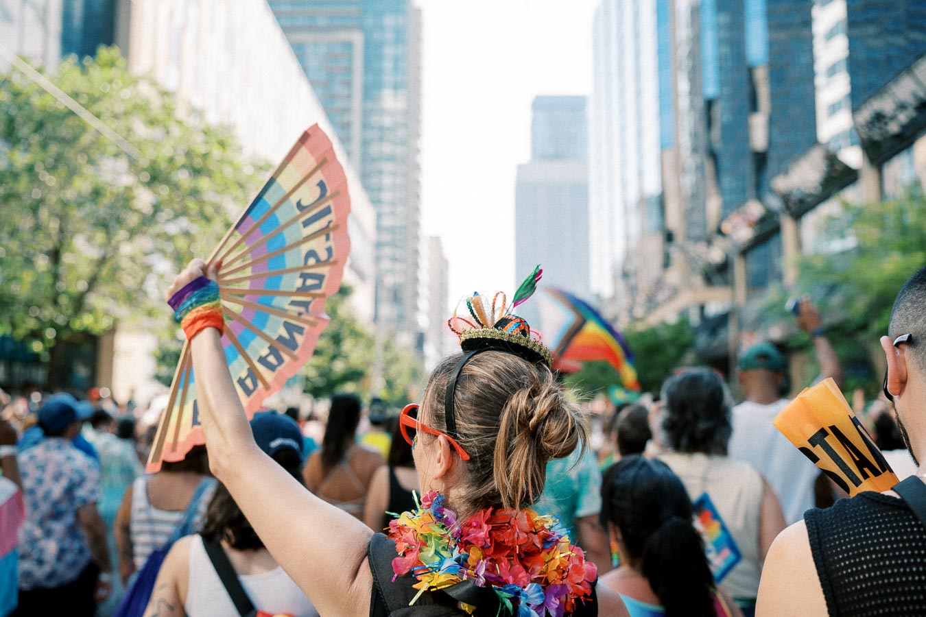 A vibrant scene at a pride parade, featuring a person with a rainbow fan and colorful accessories, surrounded by a lively crowd in a city setting.