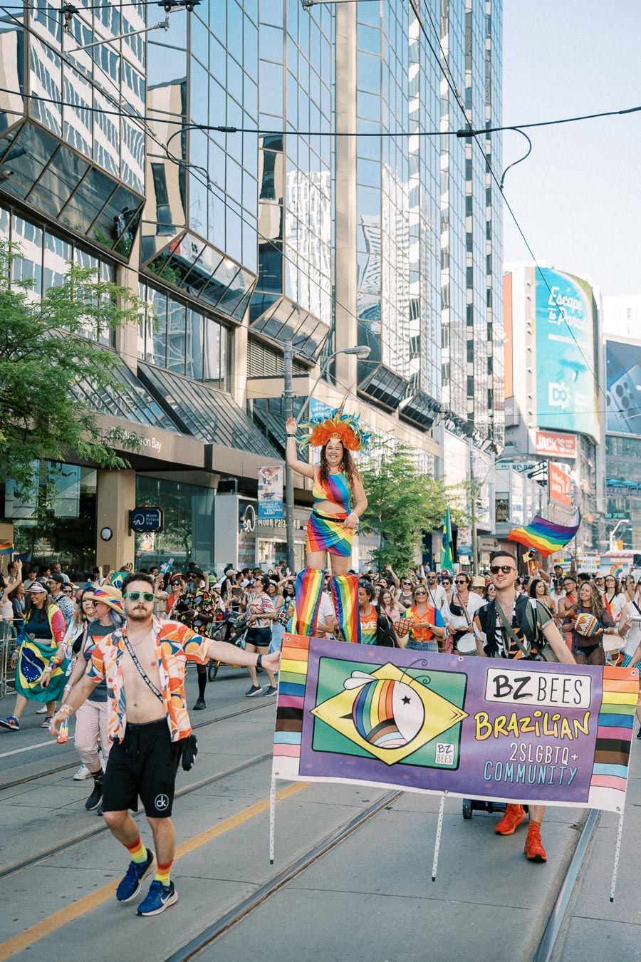 Participants in a parade celebrate with a banner for the Brazilian 2SLGBTQ+ community, showcasing colorful attire and a lively atmosphere.