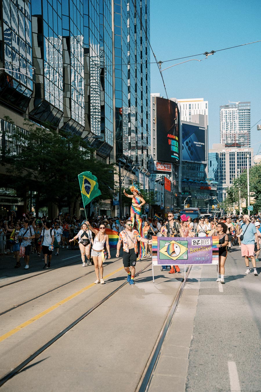 Participants in a parade march down a city street, holding a banner for the Brazilian LGBTQ+ community. The scene is lively with colorful outfits and flags, set against a backdrop of tall buildings.