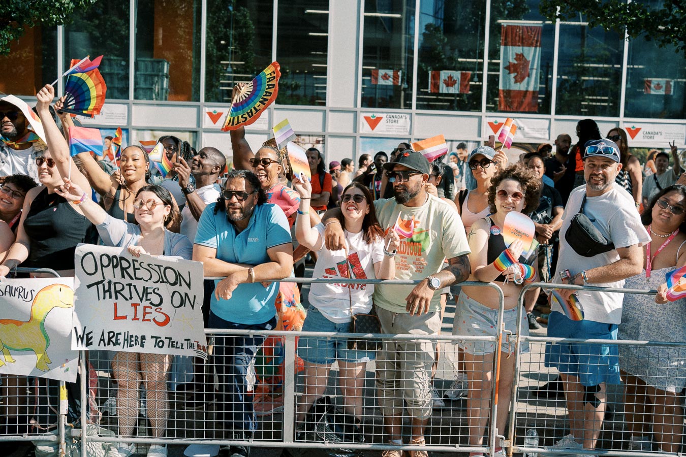 A lively crowd celebrates at a pride parade, showcasing diversity and support with rainbow flags and signs. Canadian flags are visible in the background, indicating the location.