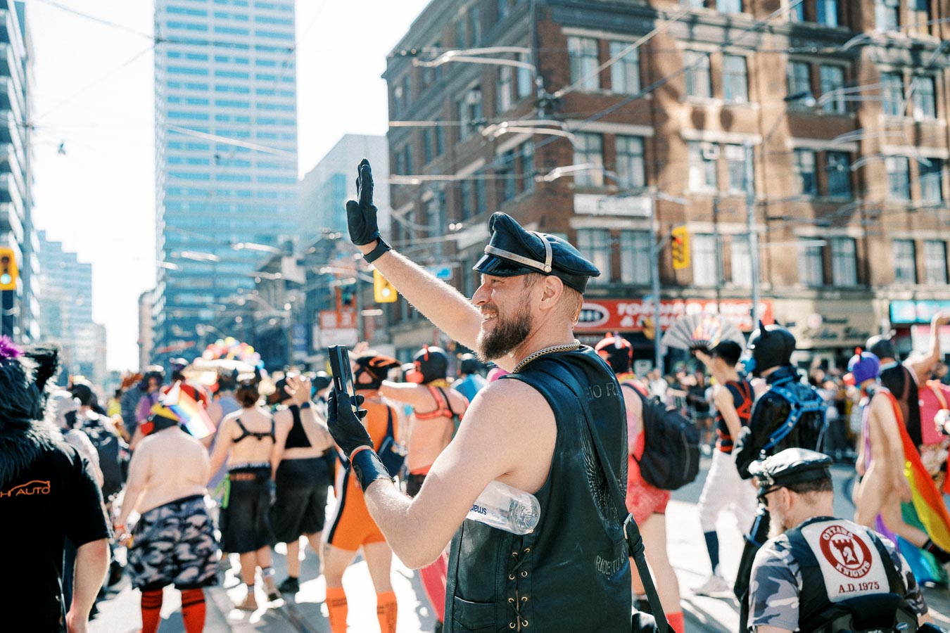 A lively parade scene with participants in colorful and diverse outfits, featuring a person in leather attire waving to the crowd.