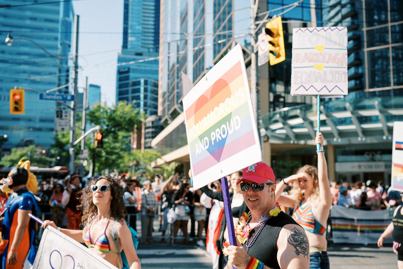 Participants in a pride parade hold signs advocating for polyamory and marriage equality, surrounded by a lively crowd and urban buildings.