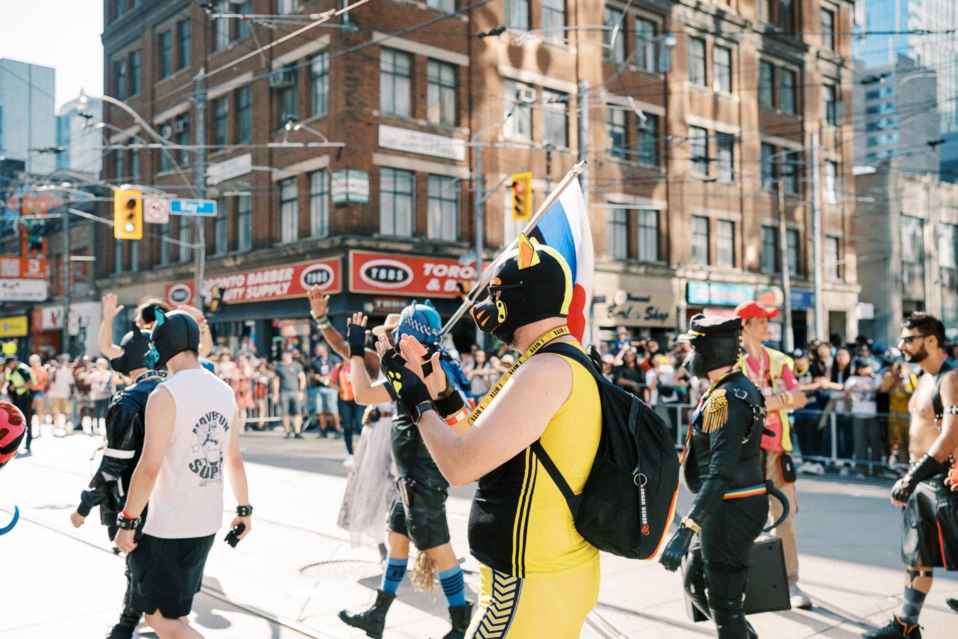 Participants in colorful costumes and masks march in a lively parade on a city street, with onlookers gathered along the sidewalks.