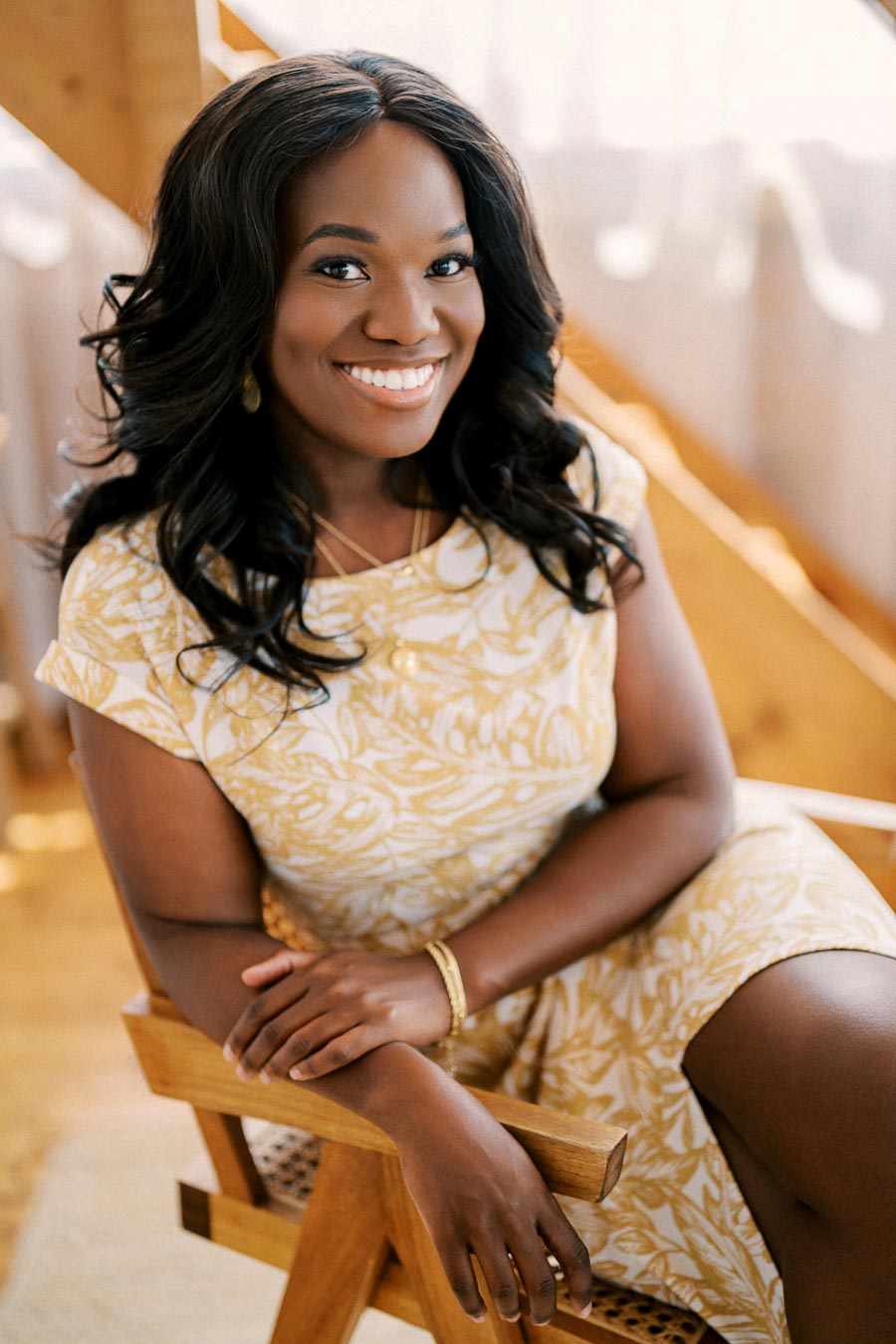 A woman in a yellow and white patterned dress smiles while sitting on a wooden chair.