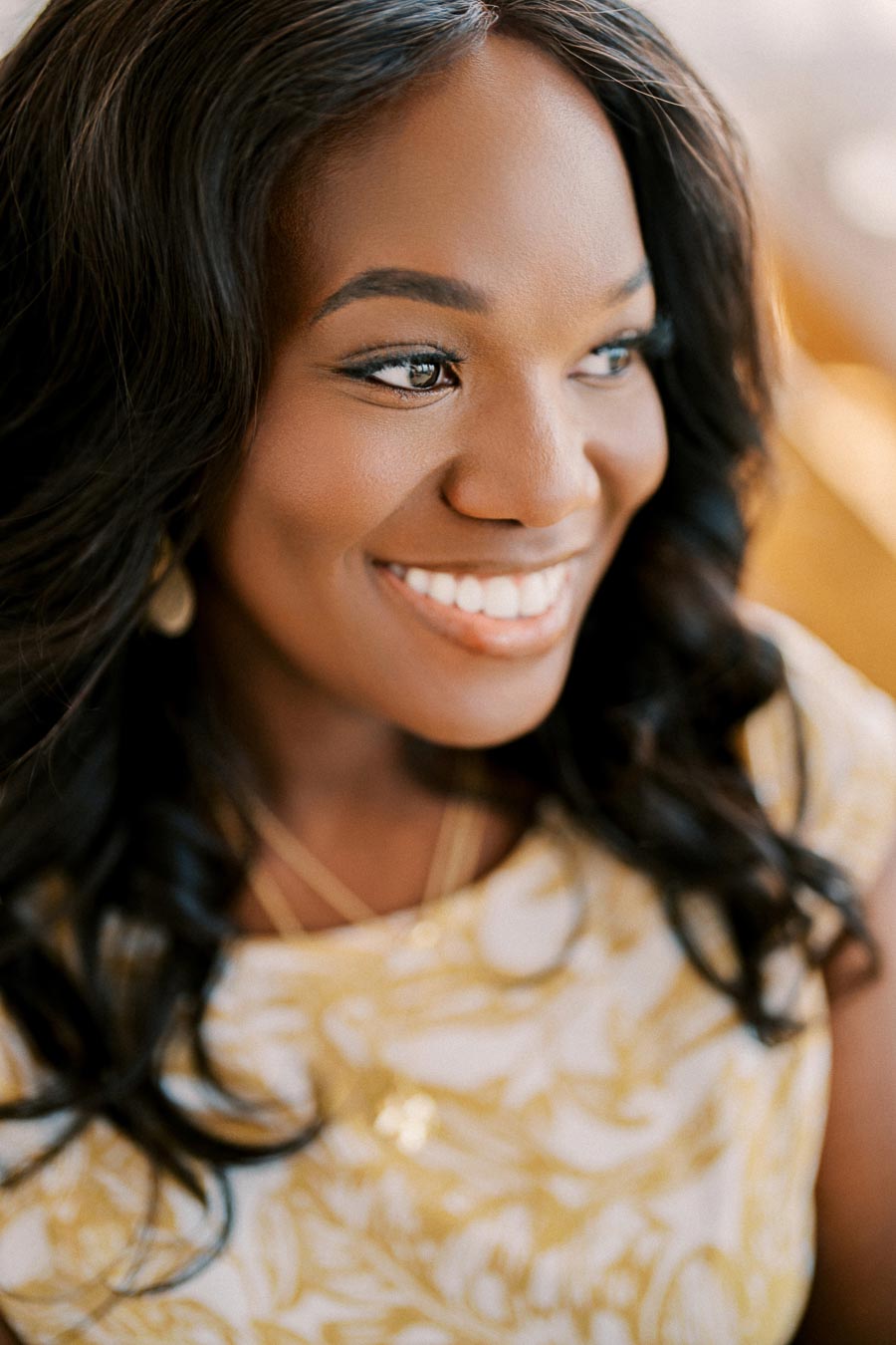 A woman smiling warmly, dressed in a white and yellow patterned dress with gold jewelry.