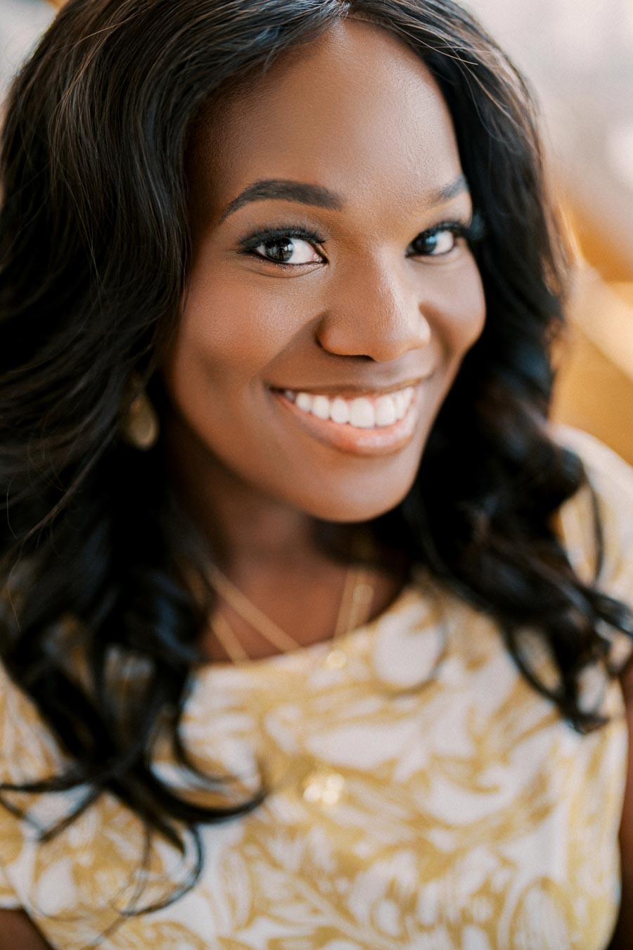 A woman with long dark hair smiles warmly, wearing a white and gold patterned top.