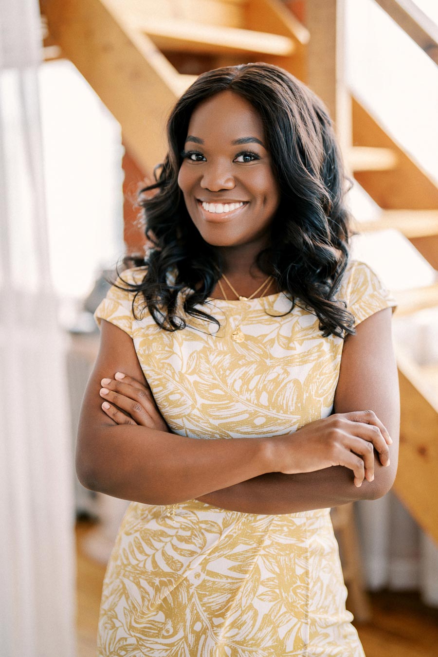 A woman smiling confidently in a yellow and white patterned dress, standing with her arms crossed.