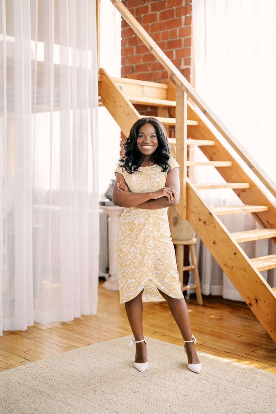A woman stands confidently in a bright room, wearing a yellow floral dress, with wooden stairs and sheer curtains in the background.