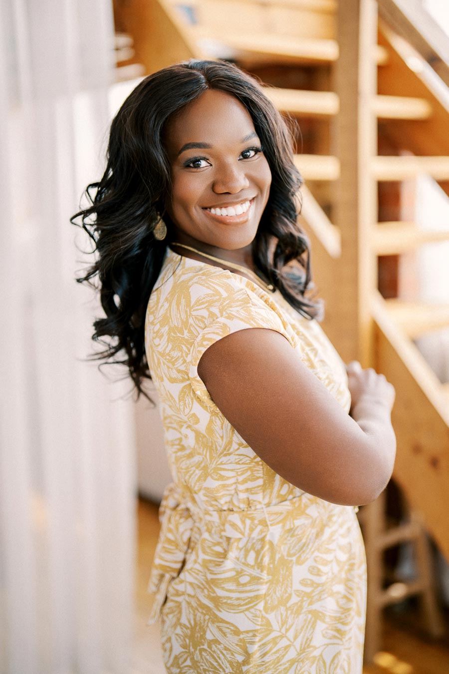 A woman in a yellow and white floral dress smiles warmly while standing indoors near a wooden staircase.