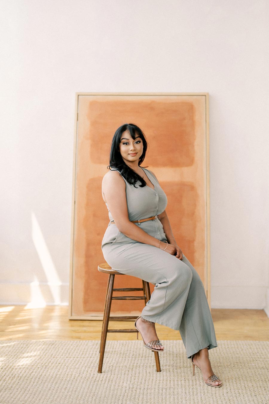 A woman in a gray jumpsuit sits on a wooden stool in a minimalist room, with a large abstract painting behind her.