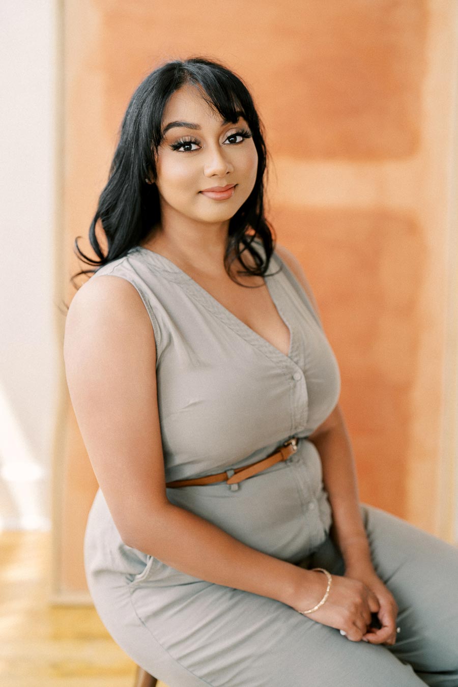 A woman with long black hair sits on a stool, wearing a gray sleeveless jumpsuit, against a warm-toned background.
