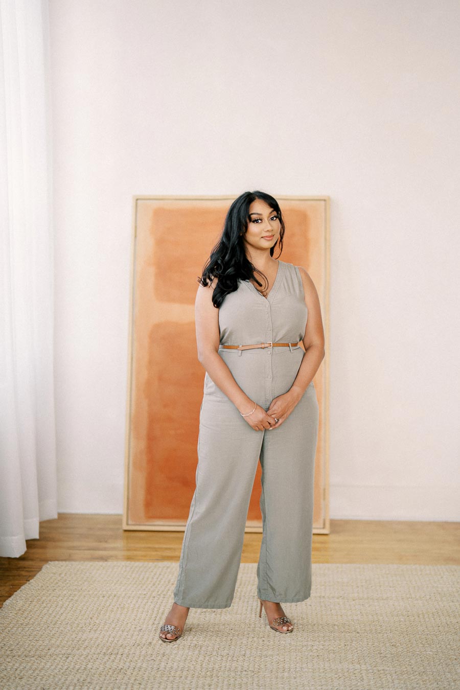 A woman stands confidently in a beige jumpsuit, posing in a minimalist room with a textured rug and a large framed artwork in the background.