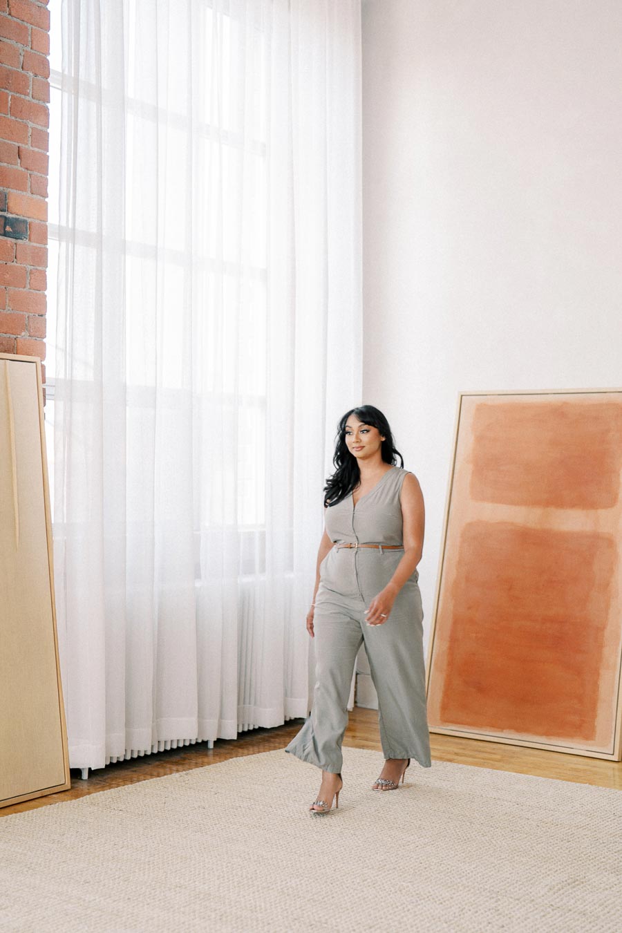 A woman in a beige jumpsuit walks in a well-lit room with large windows and abstract art pieces leaning against the wall.