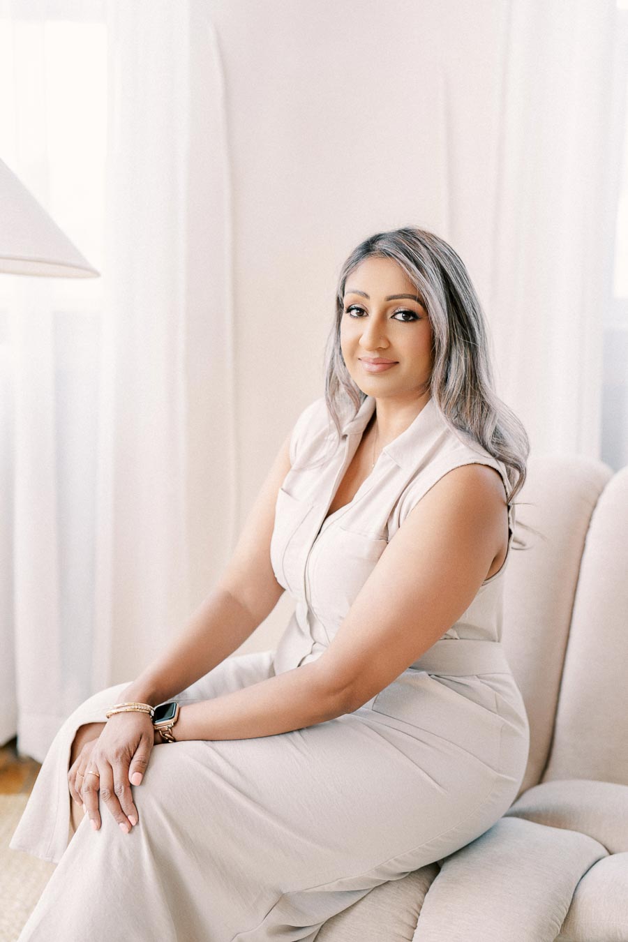 A woman sits comfortably on a beige couch, wearing a light-colored sleeveless dress and a smartwatch, with a soft smile and relaxed posture.