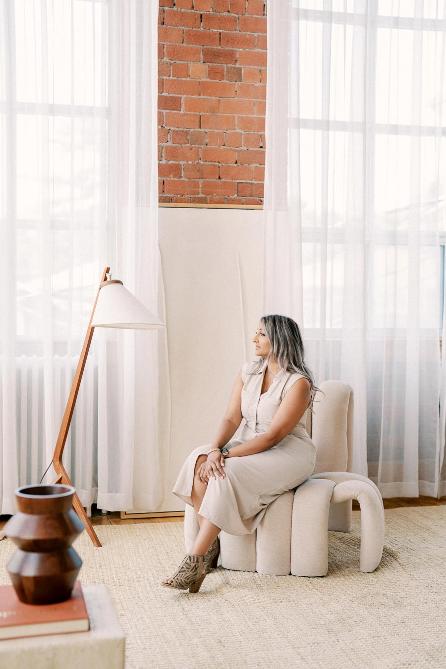 A woman sits on a modern chair in a stylish room with brick walls and sheer curtains, next to a wooden floor lamp.
