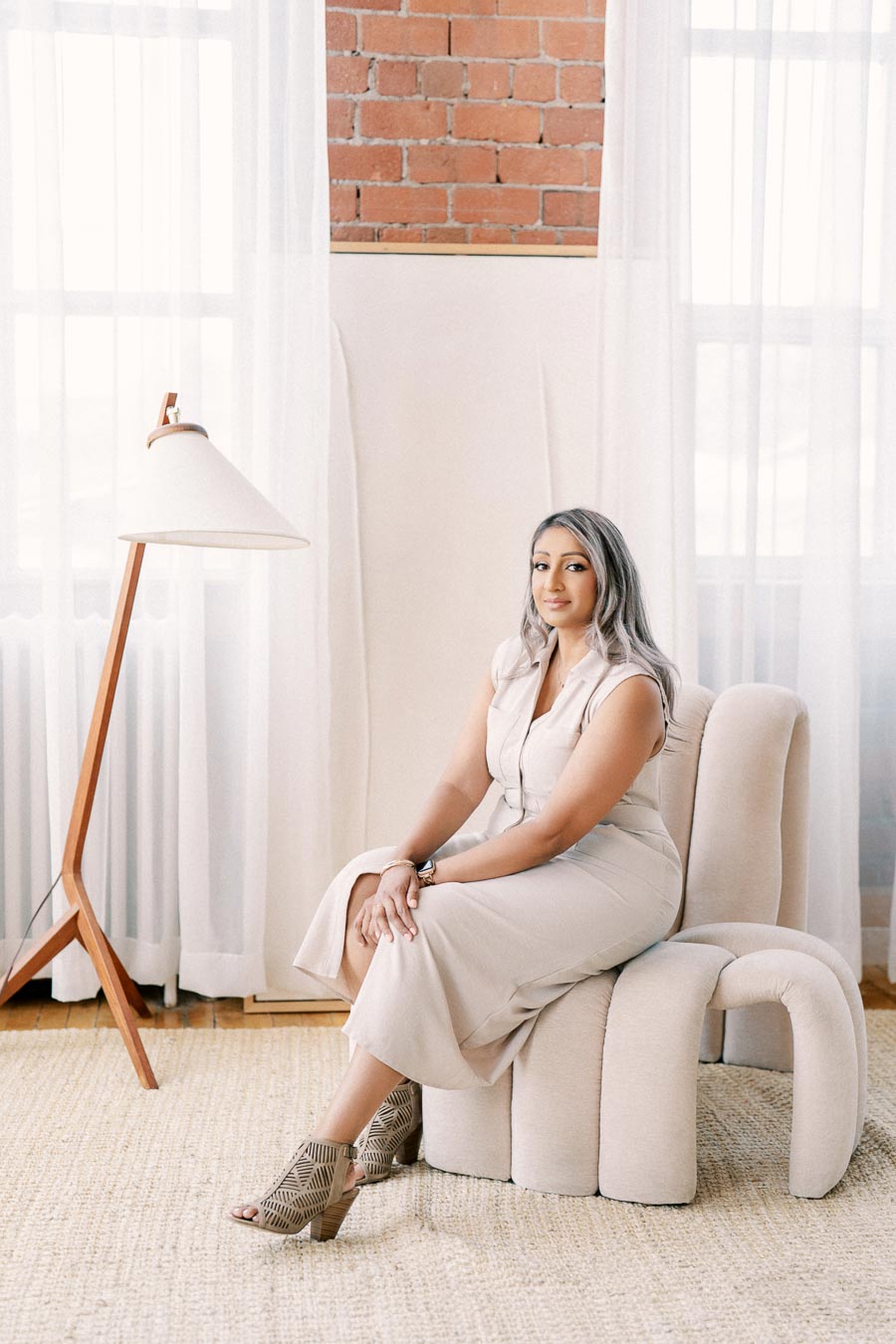 A woman in a beige dress sits on a modern chair in a stylish room with a brick wall and a wooden floor lamp.