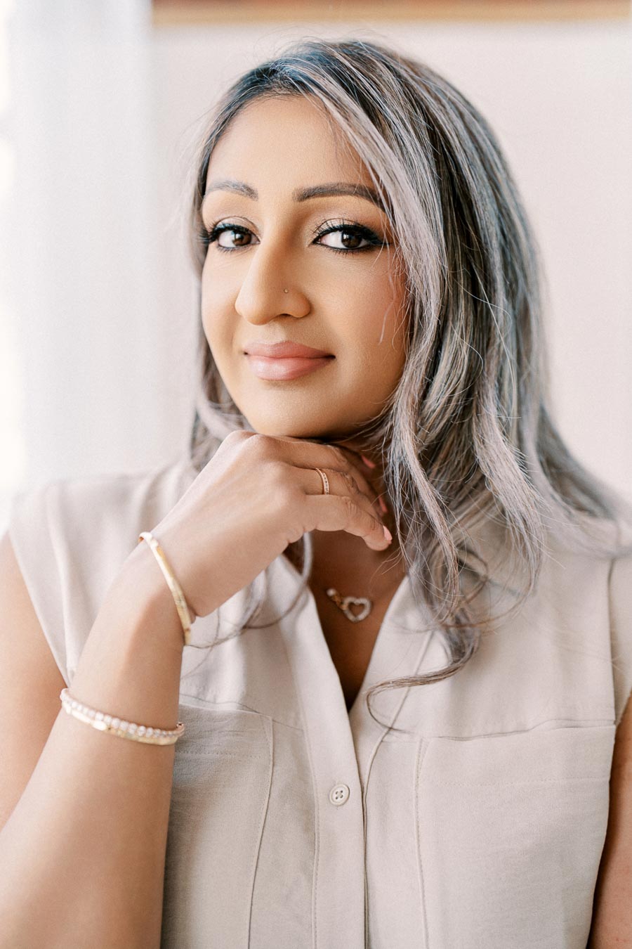 A woman poses with her hand under her chin, wearing a beige top and jewelry.