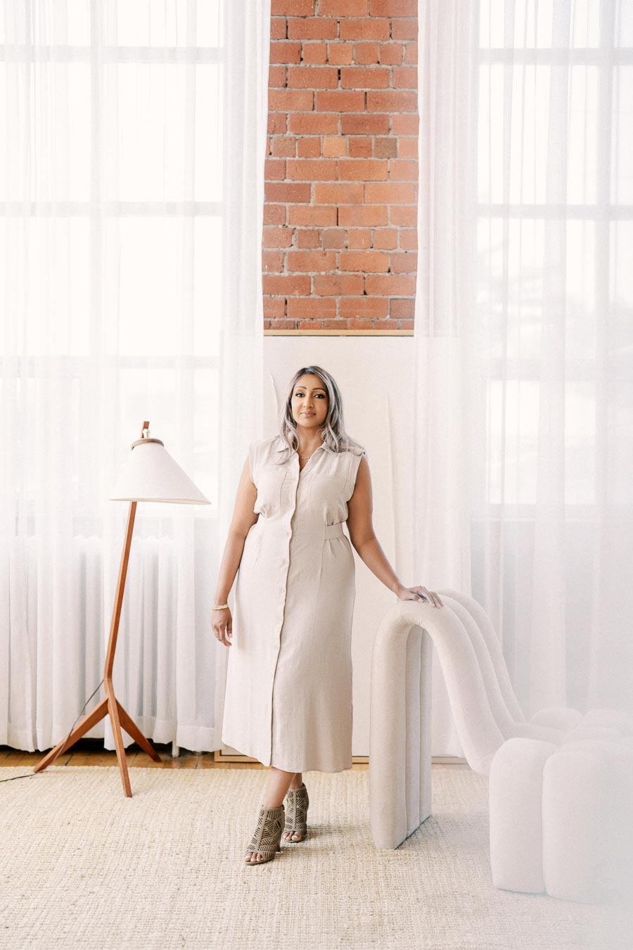 A woman stands in a stylish room featuring a brick wall, sheer white curtains, a modern floor lamp, and a uniquely designed chair.