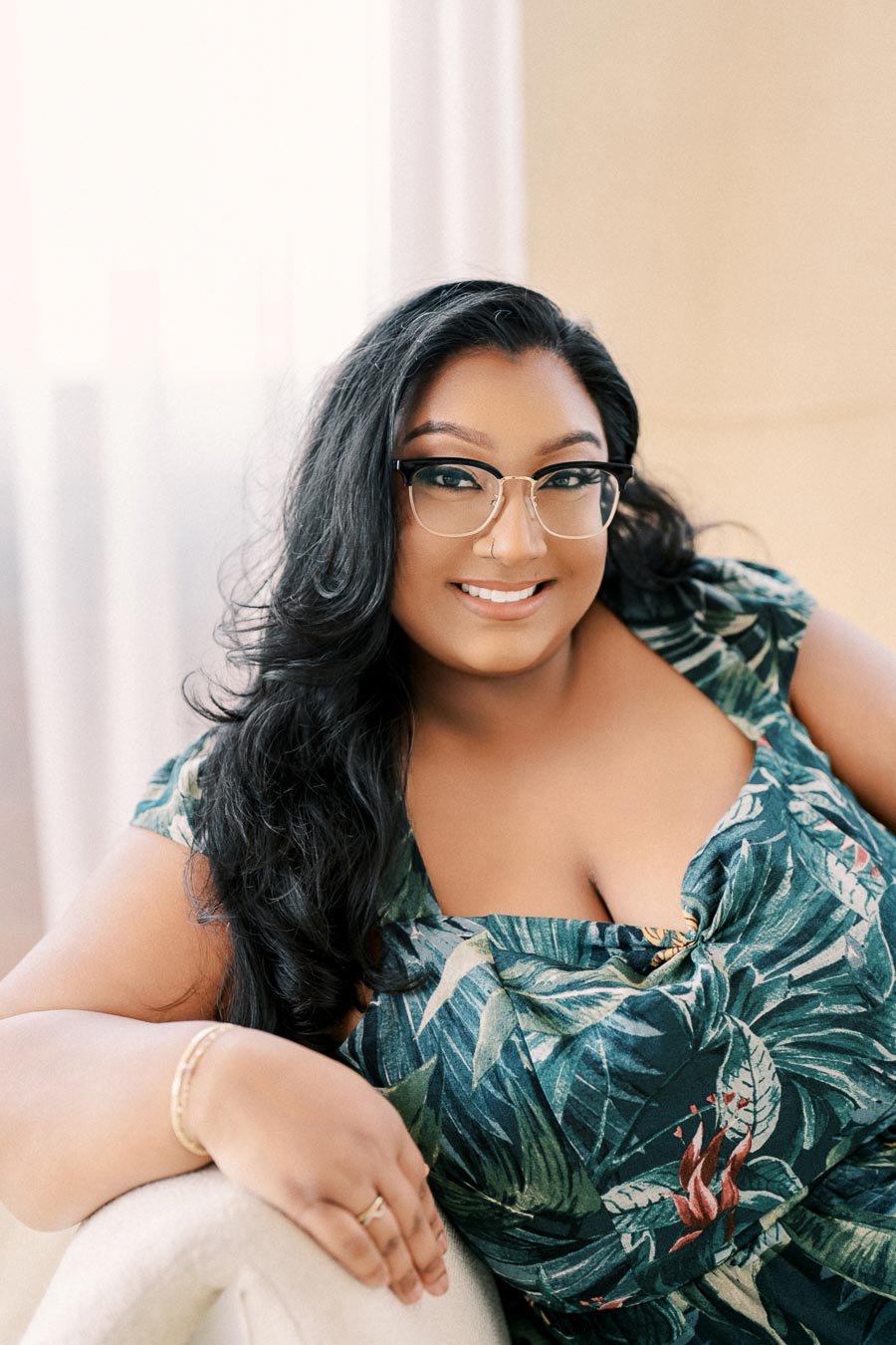 A person smiling while wearing a green tropical print dress and glasses, leaning on a white chair.