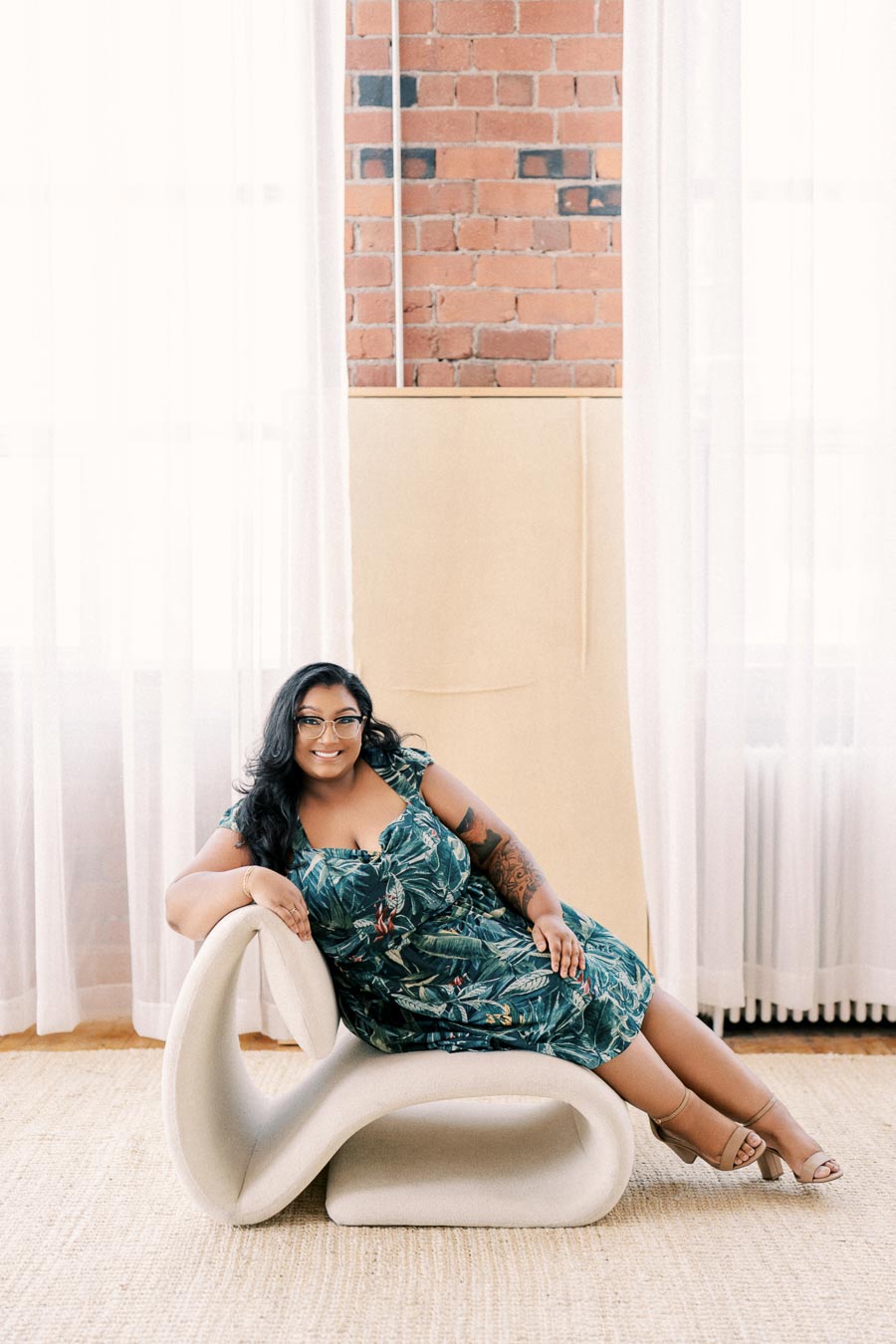 A woman in a green floral dress sits on a modern white chair, set against a backdrop of brick walls and white curtains.
