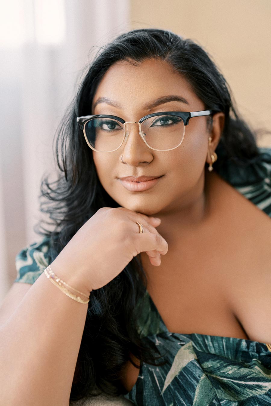 A woman with glasses and long black hair poses confidently, wearing a green patterned top.