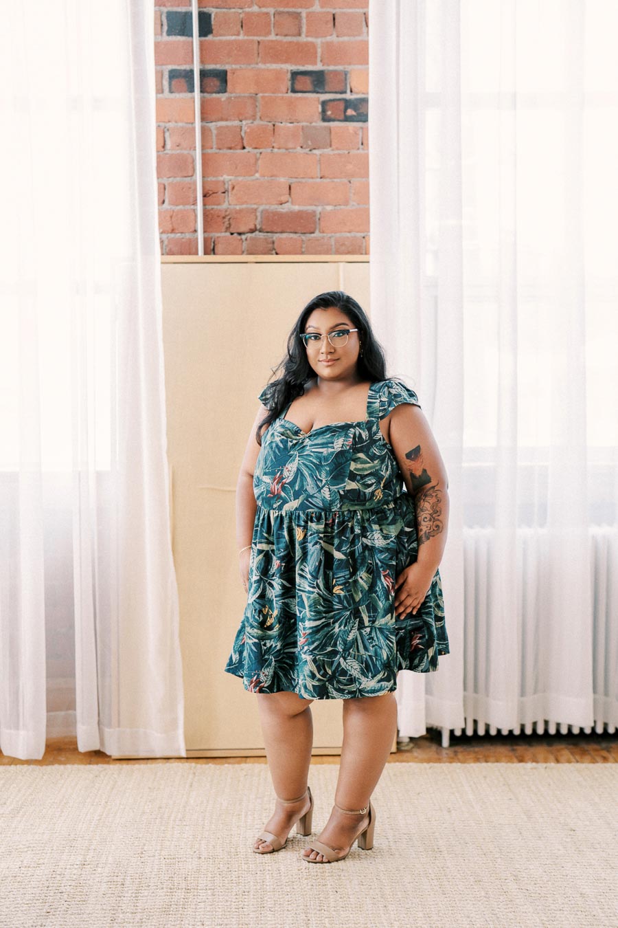 A person stands confidently in a room with brick walls, wearing a green floral dress and glasses.