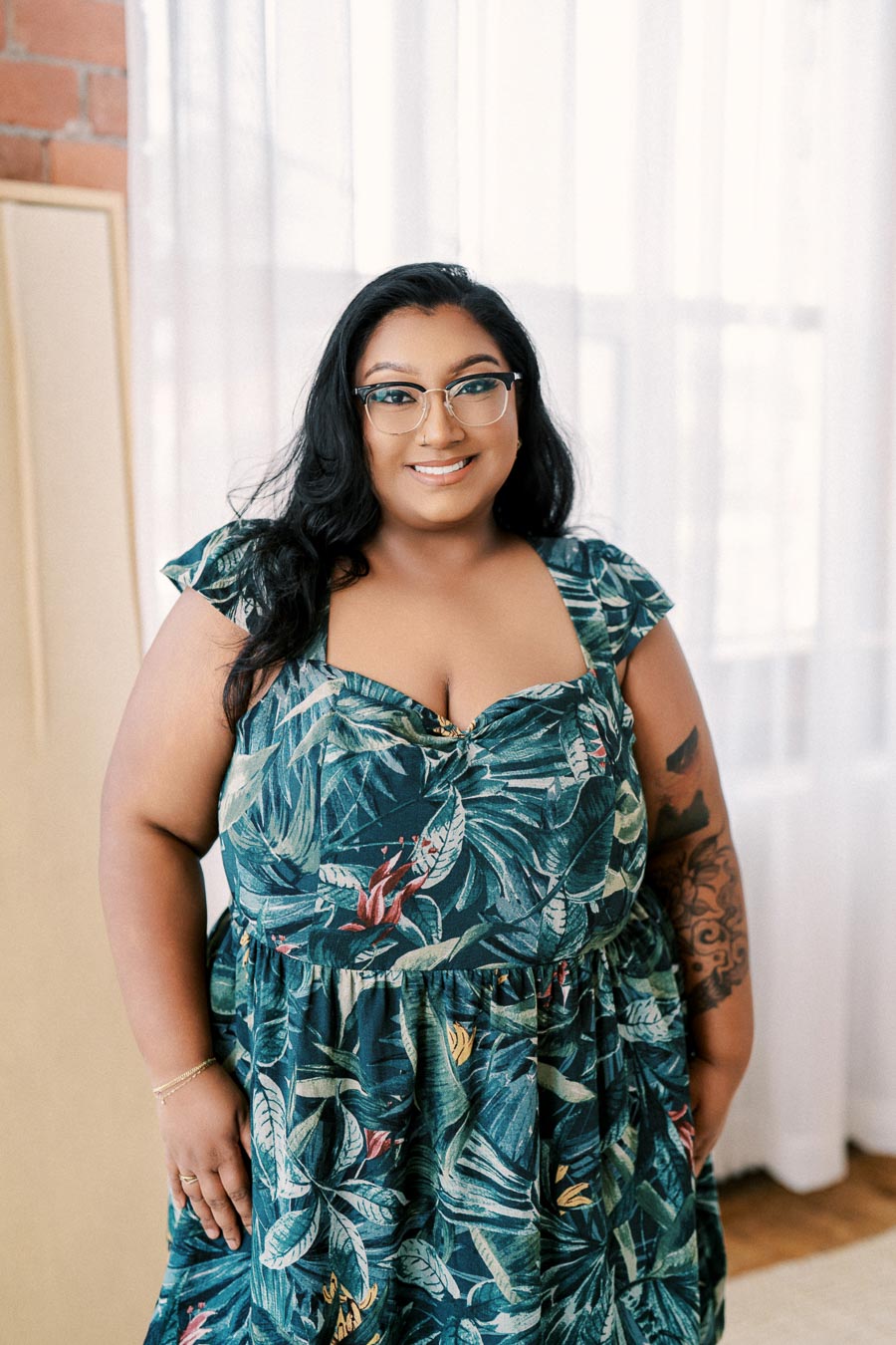 A person smiling indoors, wearing a tropical print dress and glasses.
