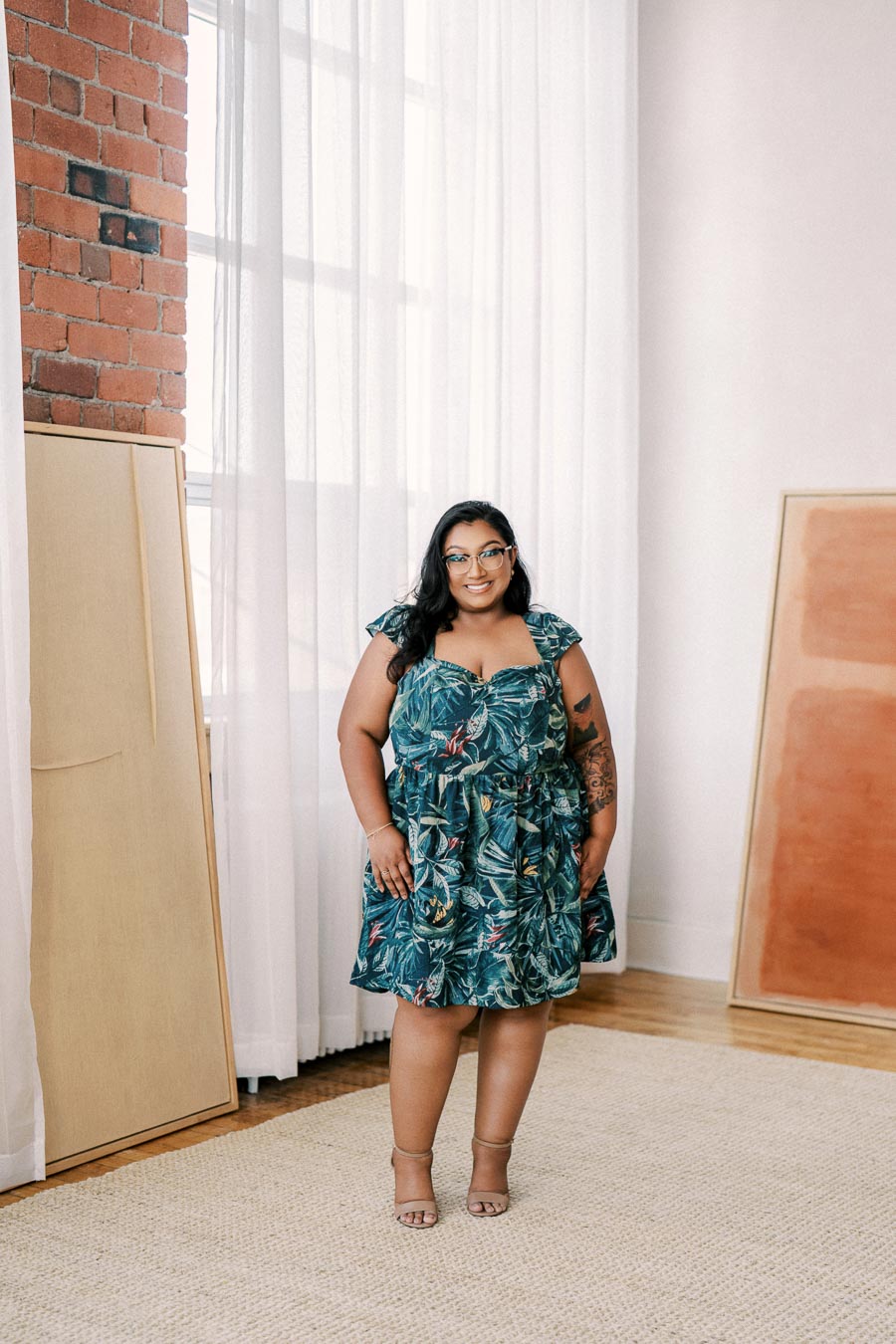 A person in a green floral dress poses in a well-lit room with white curtains and brick walls, surrounded by framed artwork.
