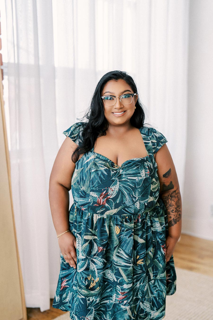 A person smiles while wearing a tropical print dress, standing indoors against a white curtain backdrop.